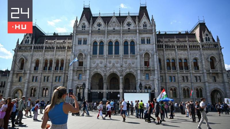A Kossuth téren egyre többen gyűlnek össze, a légkör feszültséggel teli. Az emberek hangos beszélgetésekbe merülnek, miközben a történésekre várakozva figyelik a körülöttük zajló eseményeket. A téren színes molinók és transzparensek hirdetik a különböző n